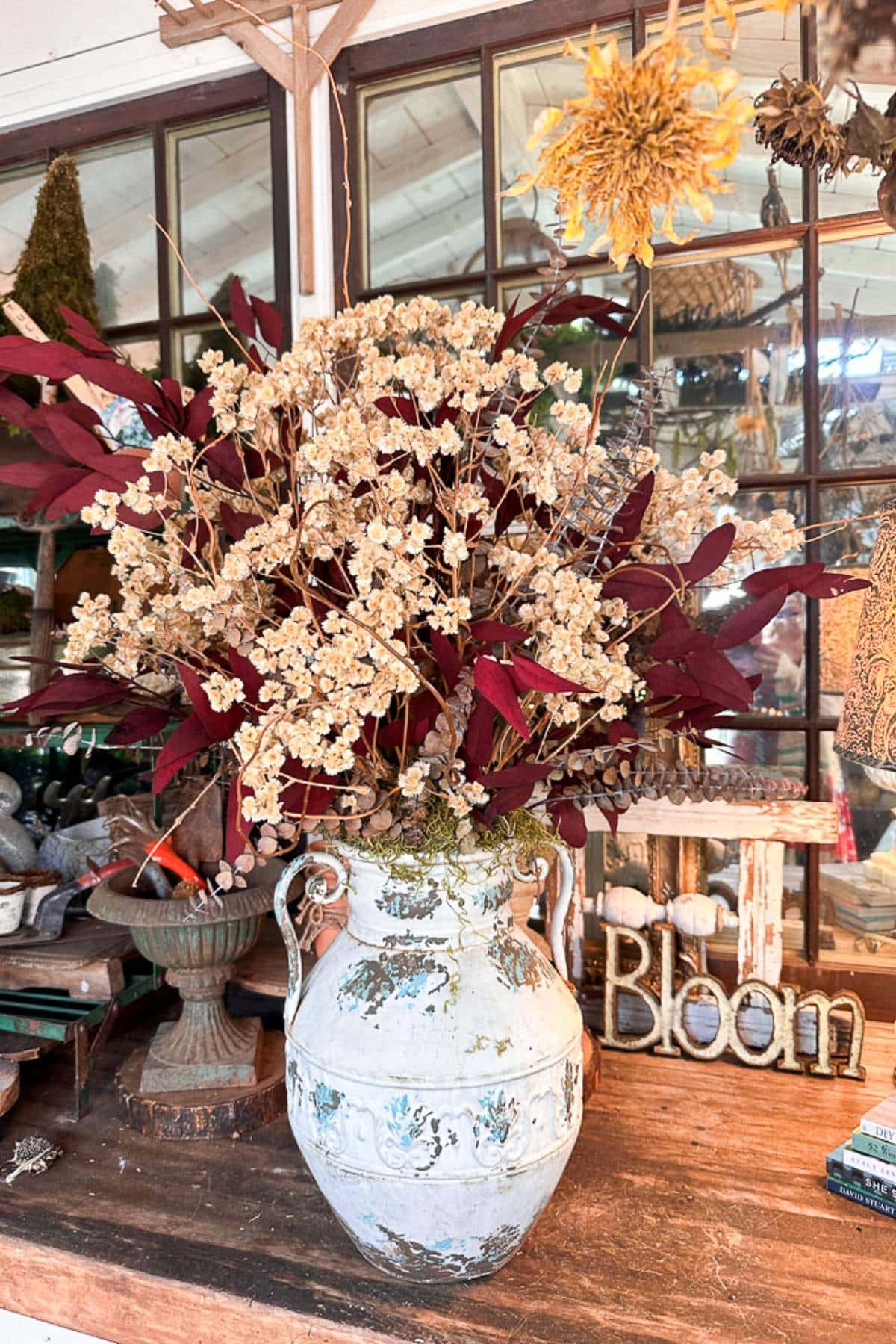 A rustic setup displays a large antique vase filled with dried flowers and foliage in shades of white and deep red. The backdrop shows a wooden table with various decorative items, including a metal sign spelling "Bloom.