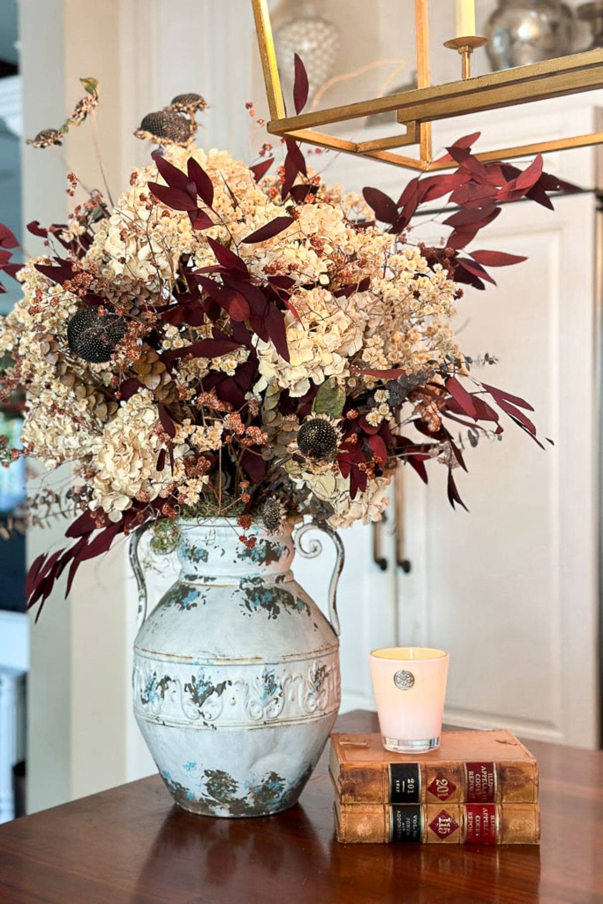 A rustic ceramic vase filled with dried flowers, including hydrangeas and red foliage, sits on a wooden table. Beside it, a lit candle rests atop two stacked vintage books with worn covers. The background features a white cabinet and a brass chandelier above.