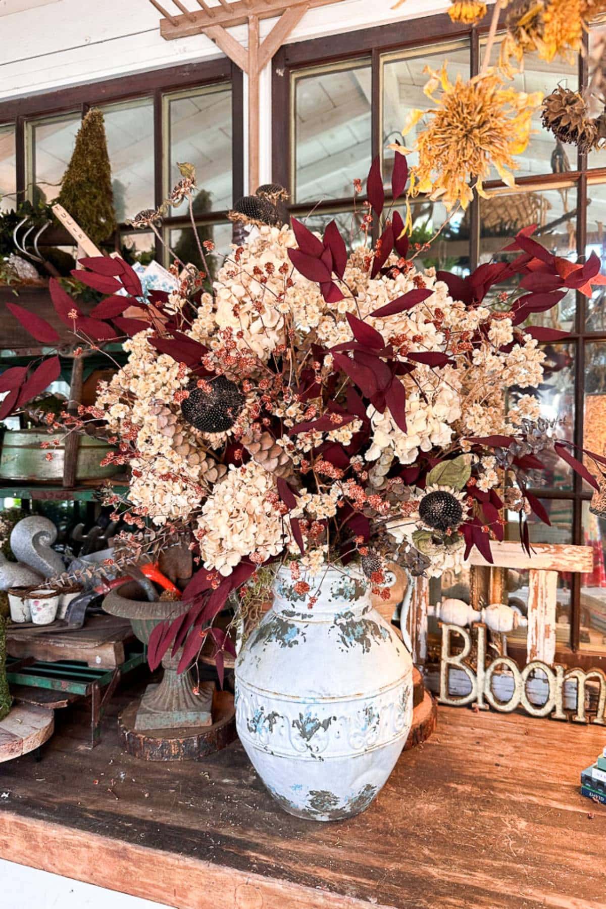 A large, ornate ceramic vase on a wooden table holds a mixed arrangement of dried flowers, including white hydrangeas and deep red foliage. In the background, there are windows and shelves displaying additional decorative items. A "Bloom" sign is visible to the right.