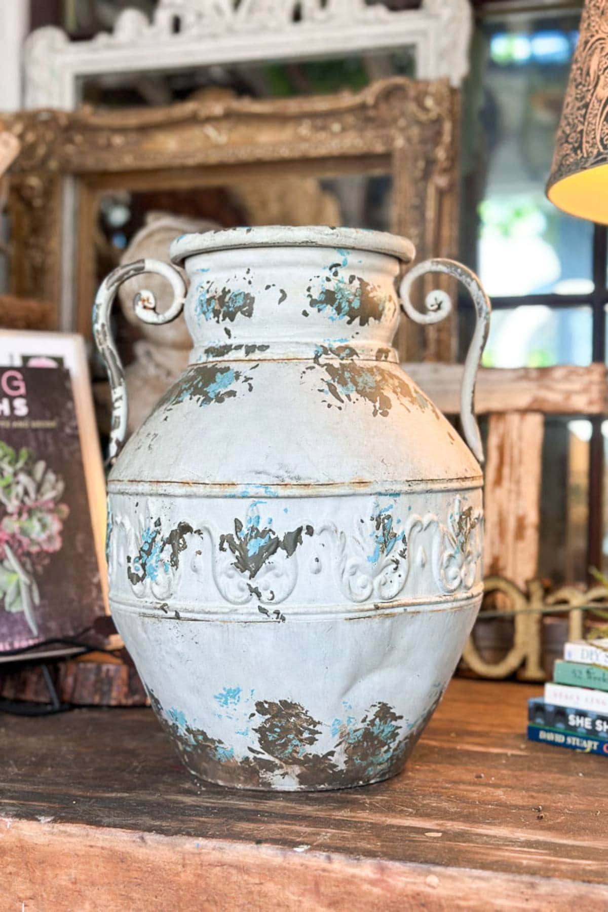 A large, weathered, white ceramic vase with blue and brown paint splotches is placed on a wooden surface. The vase features ornate detailing along its middle and two curled handles on either side. In the background, there are books, a framed mirror, and a lamp.