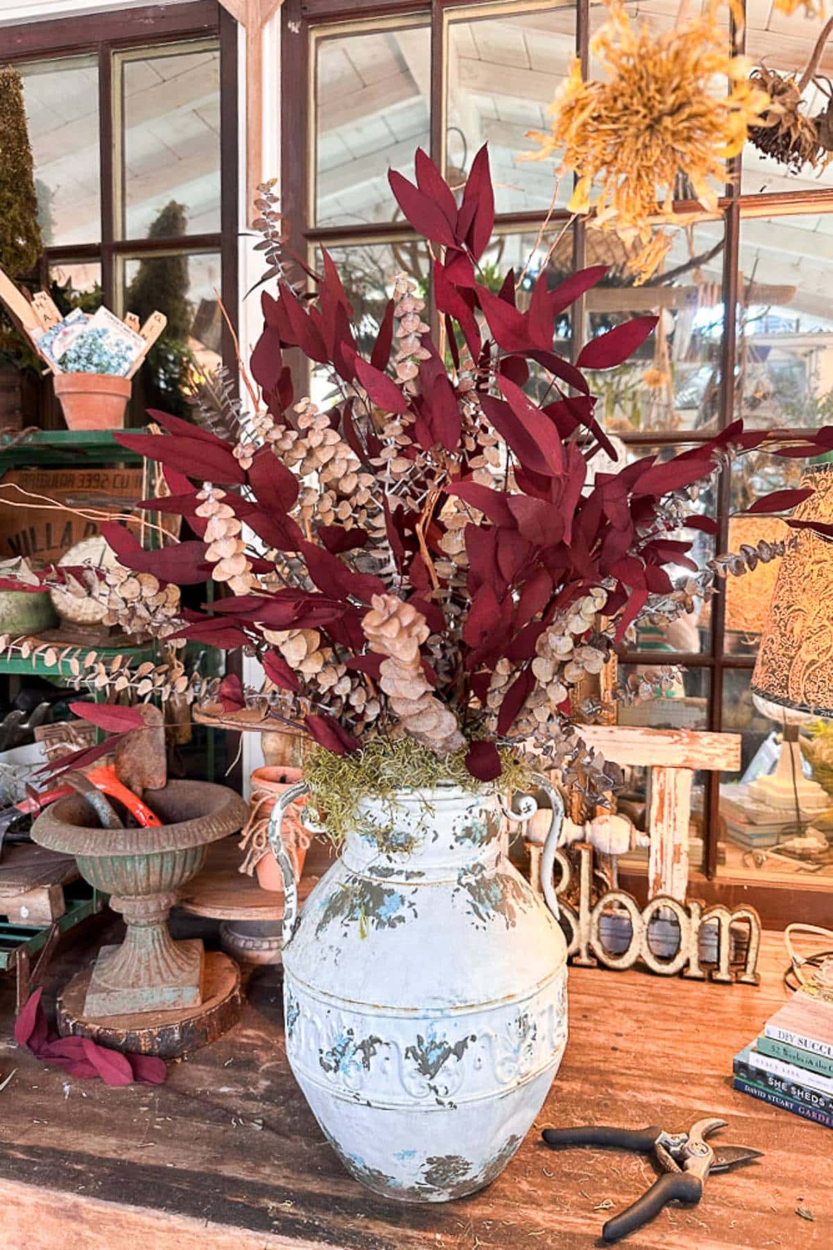 A white-painted ceramic vase with a distressed finish holds burgundy and beige dried foliage. The vase is placed on a rustic wooden table in front of a large window with multiple panes. Various garden tools and decorations are scattered around the area.