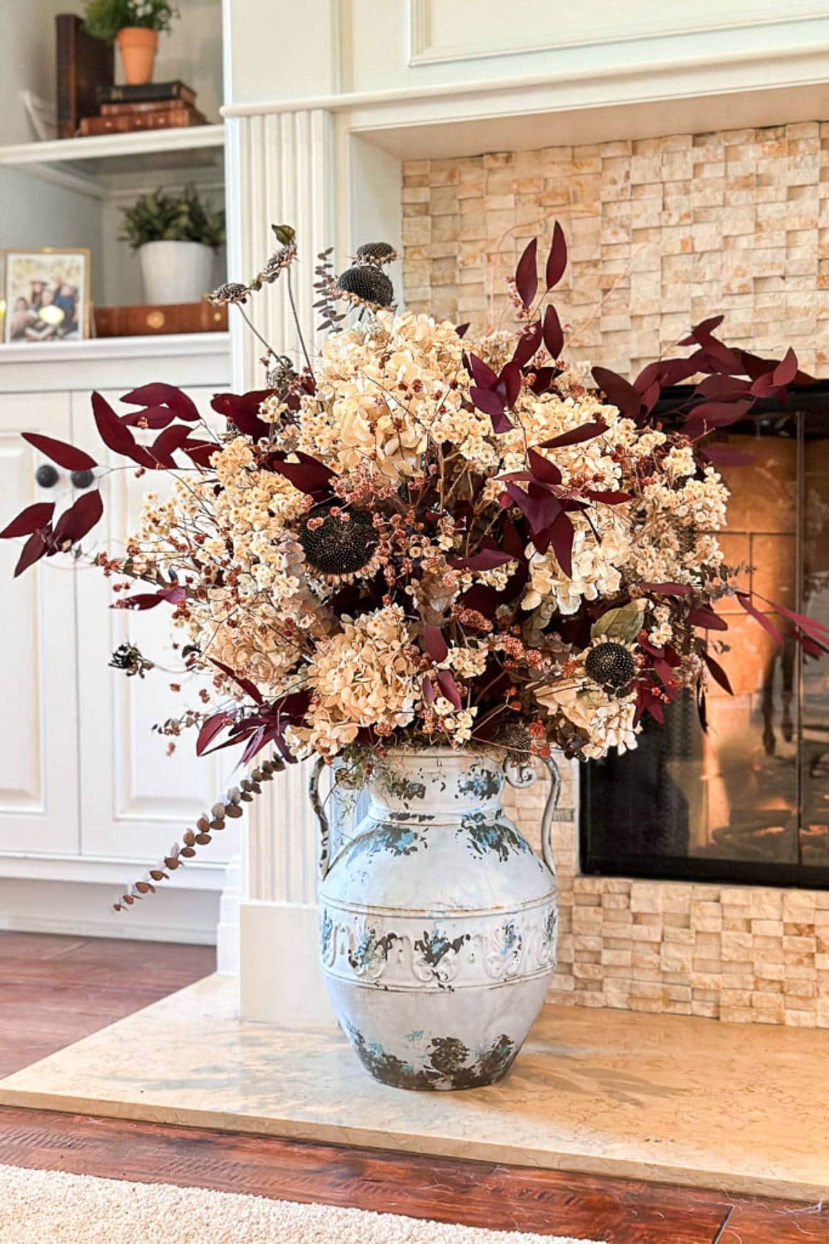 A large, ornate vase filled with a lush arrangement of dried flowers and leaves in hues of cream, burgundy, and brown stands in front of a tiled fireplace. The vase has an intricate blue and white pattern, and a bookshelf is visible in the background.