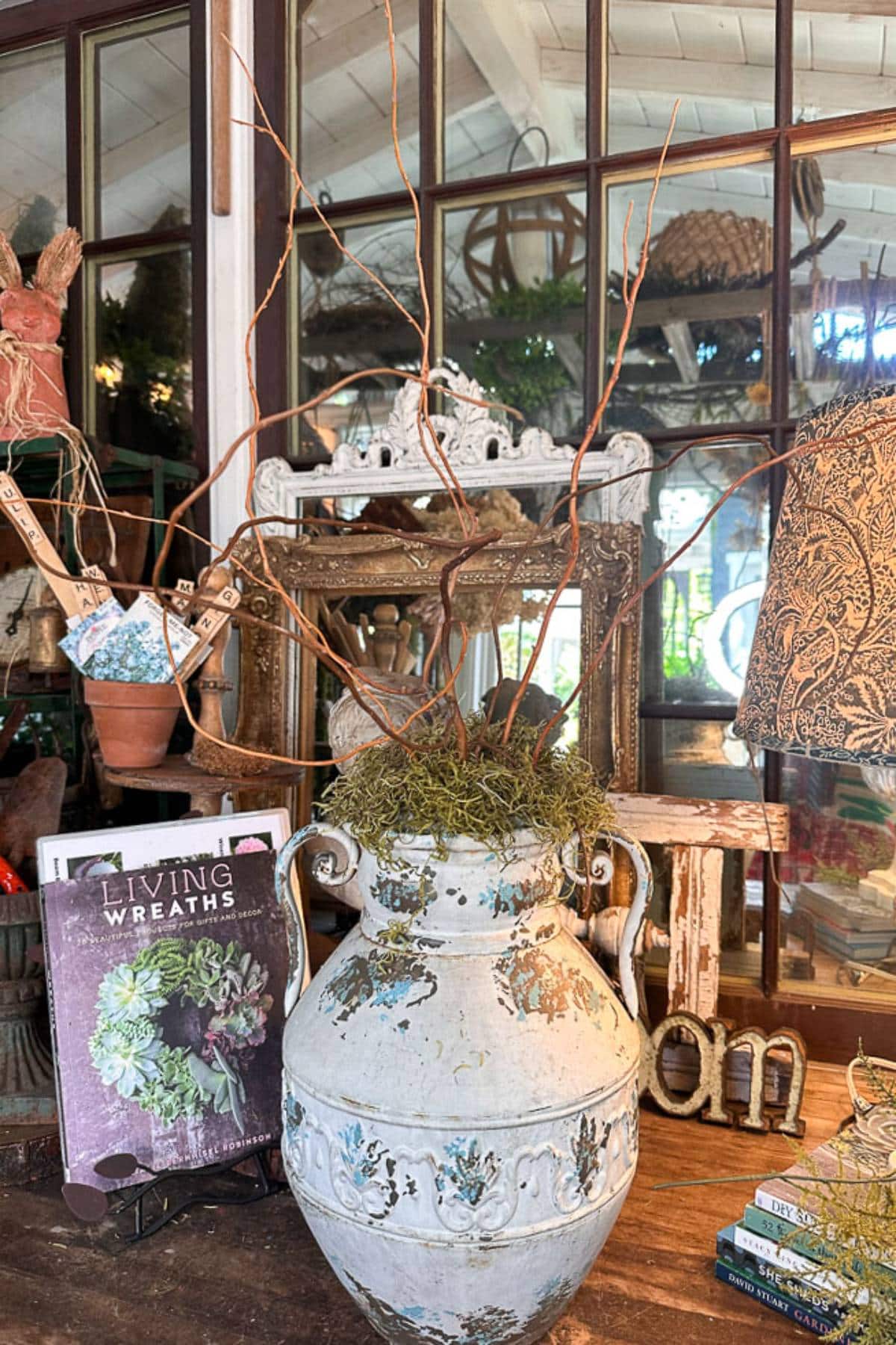 A rustic interior scene featuring a large, weathered ceramic vase filled with twigs and moss, placed on a wooden table. Surrounding the vase are various decorative items, including a book titled "Living Wreaths" and a small sign that reads "home".