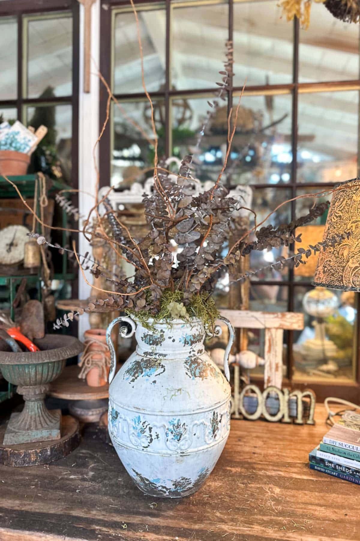 A large, decorative ceramic vase with two small handles stands on a wooden table. It is filled with dry, brown branches and eucalyptus sprigs. The background features a rustic window pane and various decorative items, including a birdbath and the word "bloom.