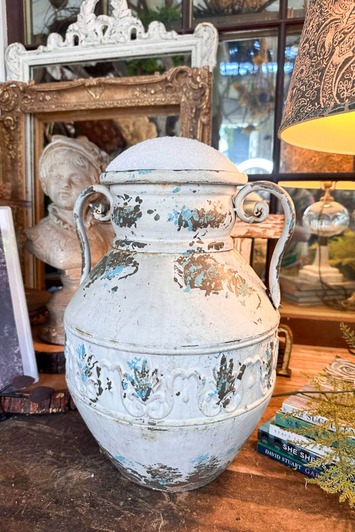 A large, weathered urn with ornate handles and faded blue and white paint sits on a wooden table. Behind it are an intricate mirror frame, a sculpted bust of a person, and a dark, patterned lampshade. Some books and other decorative items also adorn the table.