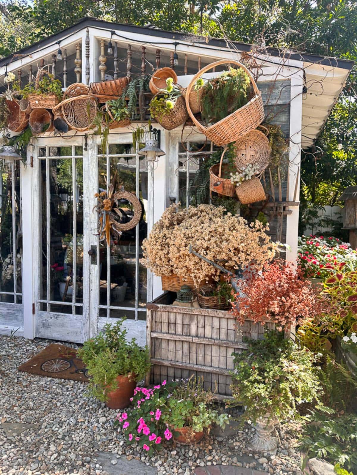 A quaint outdoor garden shop with a rustic vibe features wicker baskets and dried plants hanging outside. There are large glass doors, surrounded by potted plants, dried flowers, and lush greenery. The gravel pathway completes the charming setting.