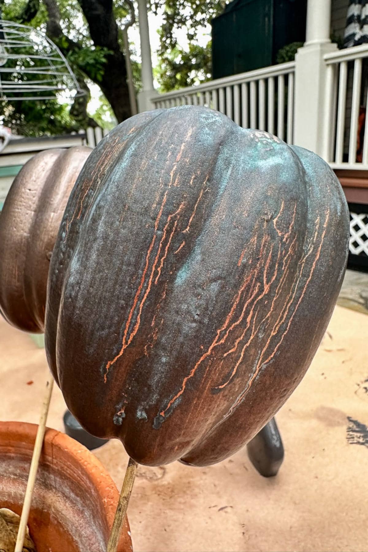 A close-up of a large, weathered, metal-look pumpkin with streaks of rust and patina serves as the perfect centerpiece for Halloween decorations. It rests on a surface with another similar pumpkin nearby, while a porch with white railings and trees creates an enchanting backdrop.