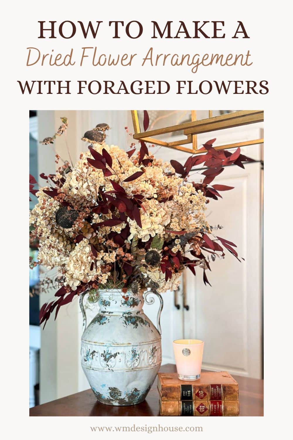 A decorative vase filled with a dried flower arrangement, featuring neutral-toned and burgundy flowers, sits on a stack of two books. A lit candle also rests on the books. The background includes a doorway. Titled "How to Make a Dried Flower Arrangement with Foraged Flowers," this image exudes charm and elegance.