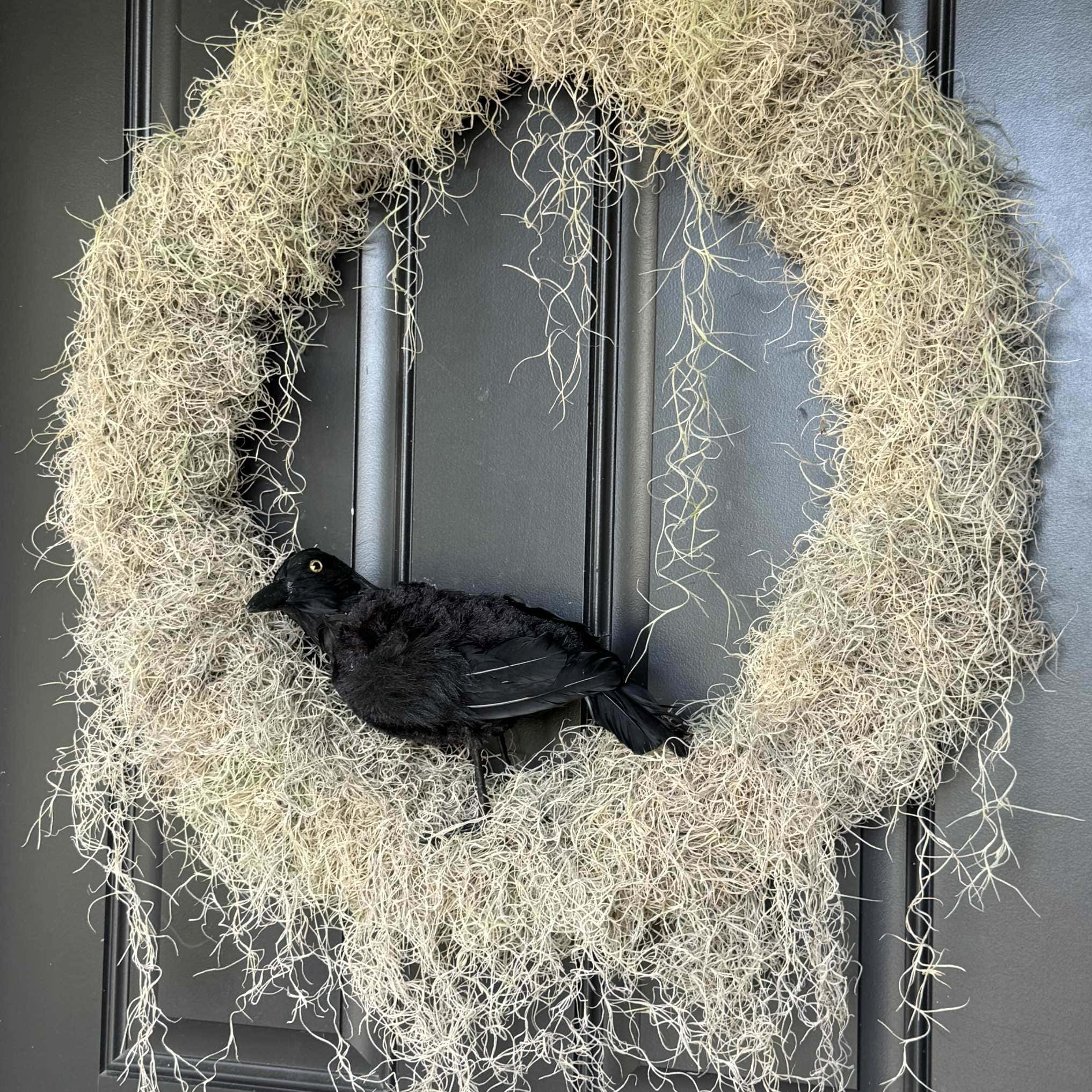 A large, circular wreath made of dried, straw-like material hangs on a dark-colored door. A realistic black crow figure is perched at the bottom of the wreath. The crow is looking to the left, contrasting starkly against the light-colored wreath. It’s labeled under HCS Code for decor in September 2024.