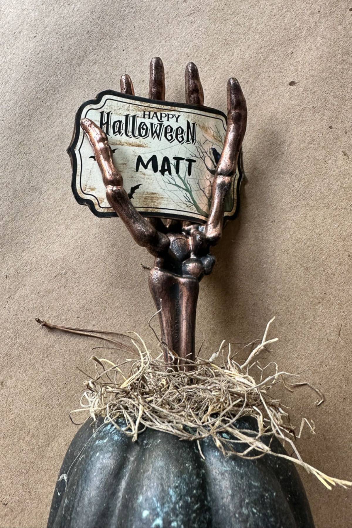 A skeletal hand emerges from a black pumpkin, clutching a sign that reads "Happy Halloween Matt" on a rustic label. Straw surrounds the pumpkin's base, crafting a spooky DIY Halloween place card holder. It's the perfect centerpiece for your Halloween table against the brown backdrop.