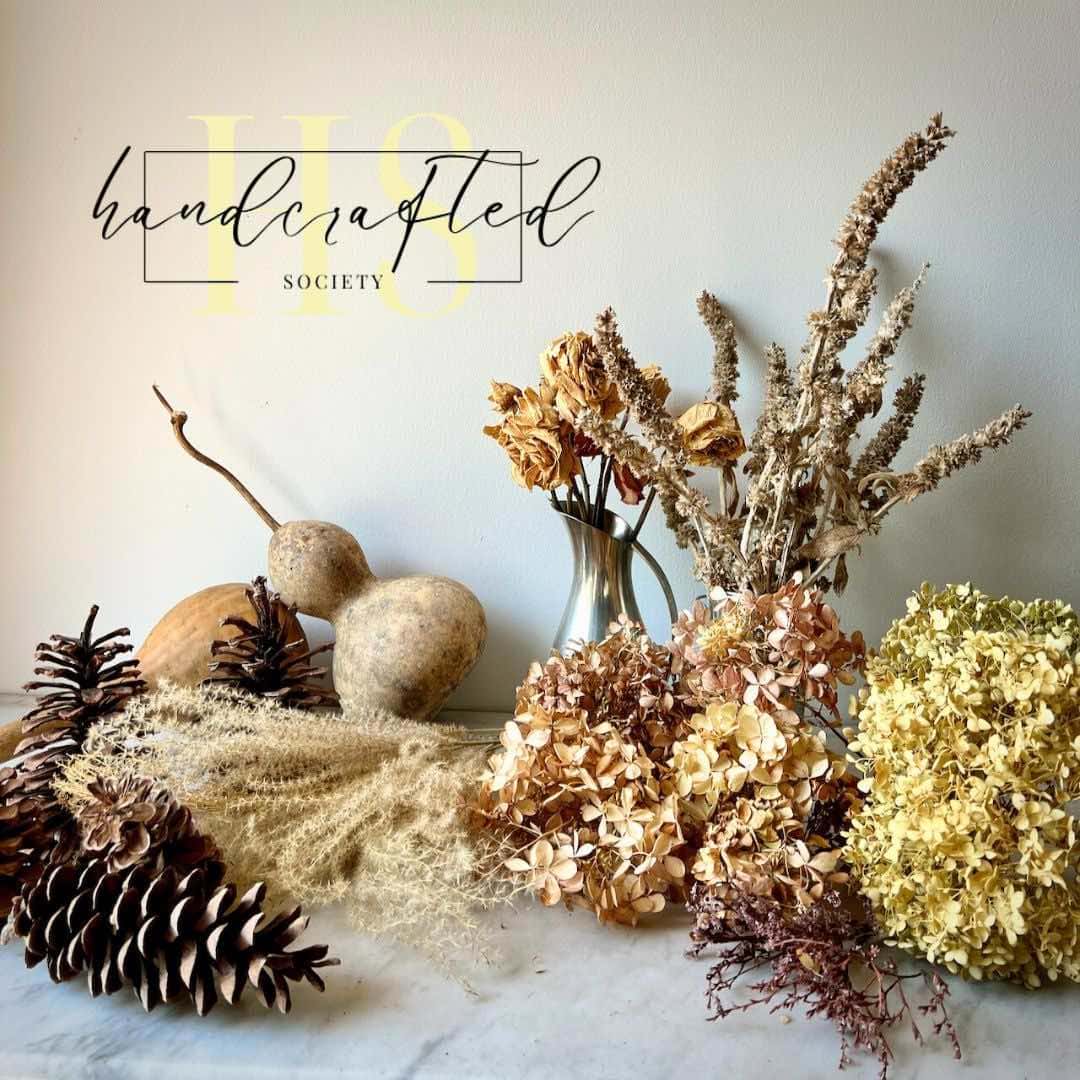 A display of various dried flowers, including hydrangeas and grasses, along with pinecones and gourds is arranged artistically on a white surface. The text "Handcrafted Society" is overlaid on the upper left corner of the image, marked with HCS Code for September 2024.