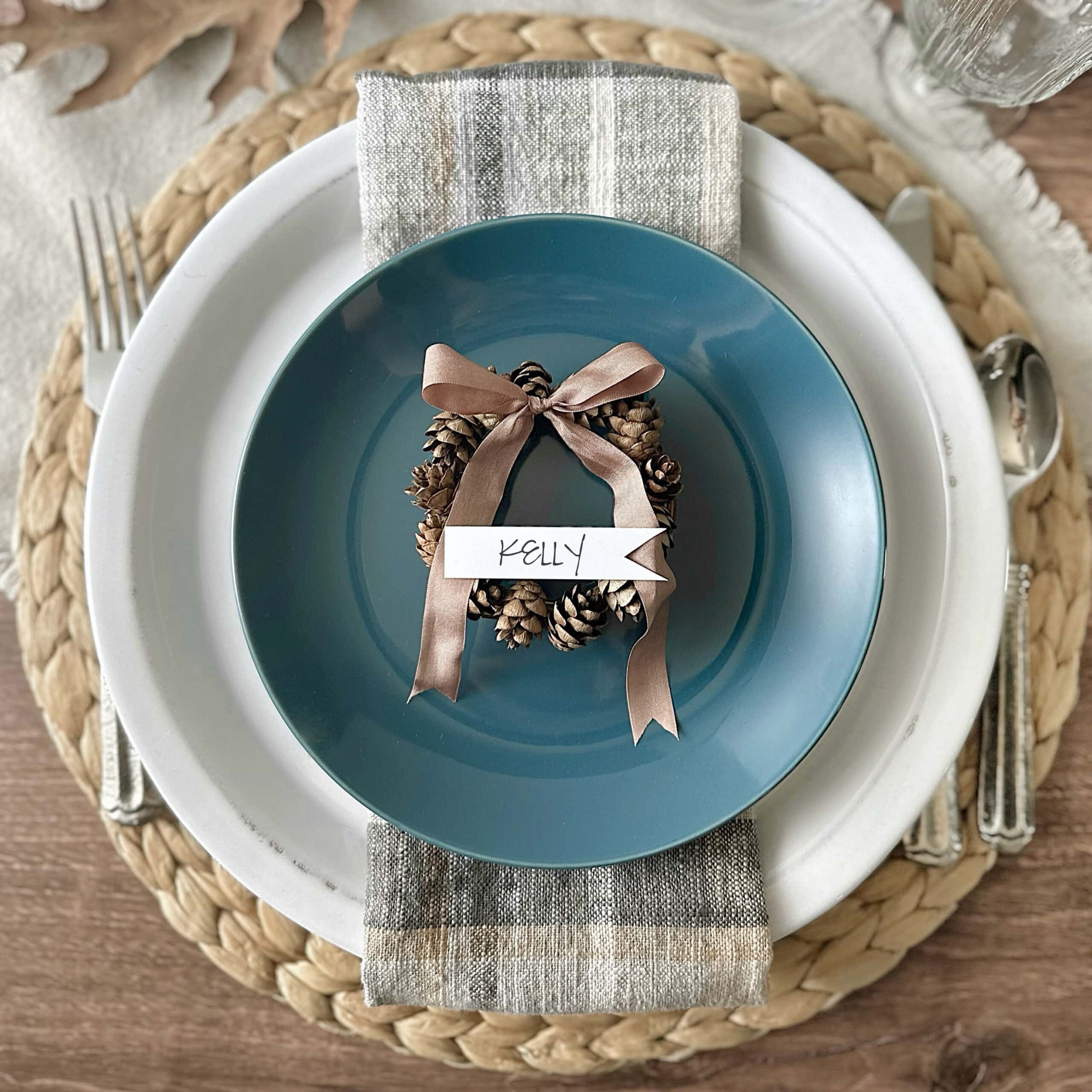 A place setting featuring a blue plate on top of a white plate with a striped fabric napkin in between. The blue plate has a small wreath made of pinecones and a brown ribbon, with a name card reading "Kelly." The setting rests on a woven placemat and a wooden table.