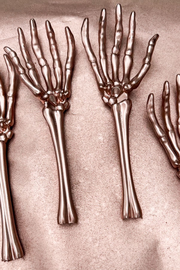 Three metallic skeleton hand forks, perfect as Halloween decorations, are arranged against a textured surface. Their elongated fingers and shiny finish make them ideal for adding a spooky touch to your Halloween table.