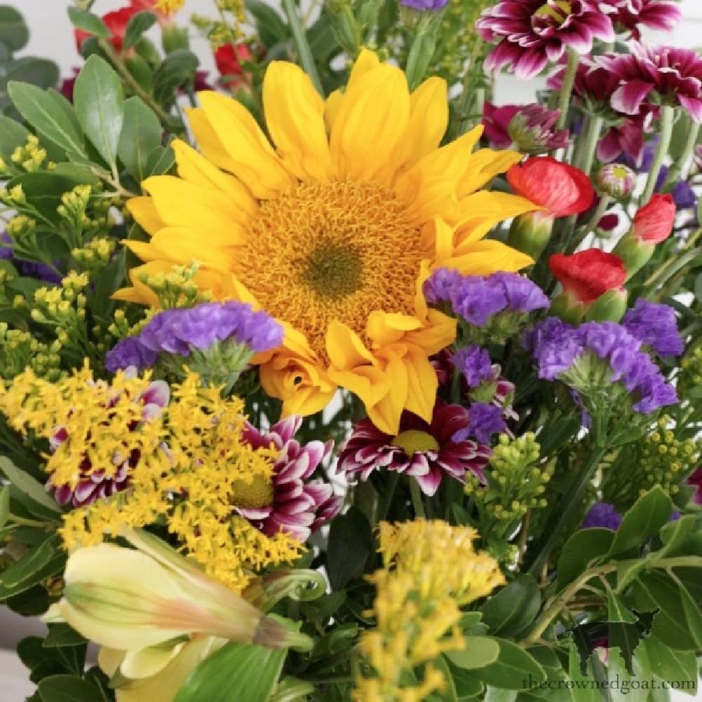 A vibrant bouquet featuring a large yellow sunflower at the center, surrounded by red and purple flowers, small yellow blooms, and lush green foliage. The mix of colors and textures creates a lively and cheerful arrangement.