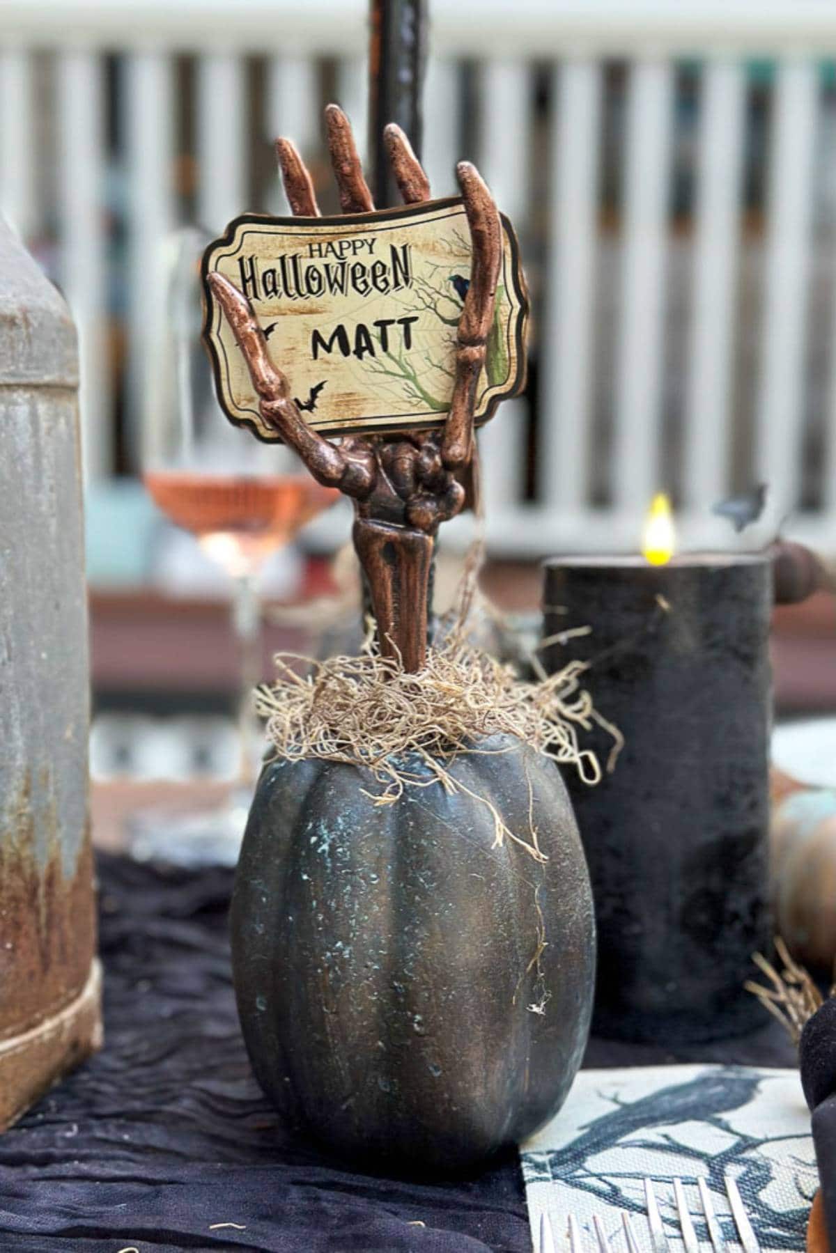 A Halloween-themed table decoration showcases a black pumpkin with a skeletal hand clutching a "Happy Halloween Matt" sign, perfect as DIY Halloween place card holders. The pumpkin is surrounded by moss, with a candle flickering in the background.