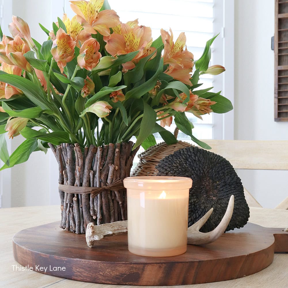 A wooden table displays a floral arrangement of orange and yellow flowers in a rustic, twig-wrapped vase. Next to it is an antler, a textured dark ball, and an ivory candle in a glass holder, all situated on a round wooden tray.