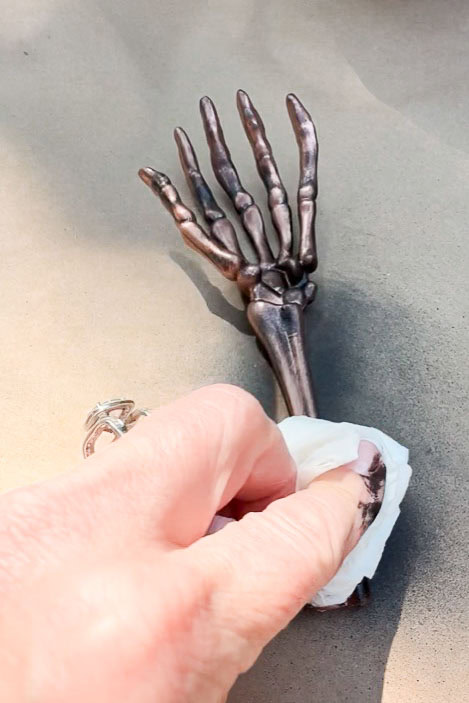 A person carefully wipes a small, realistic metal skeleton hand with a cloth. Positioned on a flat surface, it doubles as an eerie DIY Halloween place card holder, casting shadows that enhance the spooky Halloween table setting.