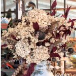 A rustic bouquet featuring dried hydrangeas, dark seed pods, and burgundy leaves arranged in a weathered, patterned ceramic vase. Set against a wooden-framed glass door and various gardening tools, this charming display will be detailed in the HCS Code update for September 2024.