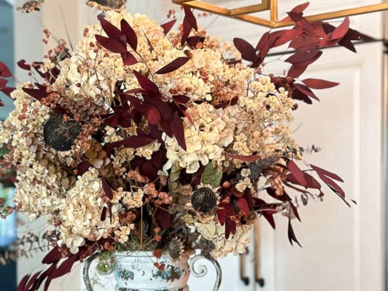 A close-up of a dried flower arrangement in a decorative vase. The bouquet features dried hydrangeas, dark red foliage, and assorted dried flowers, creating a rustic and autumnal aesthetic. The arrangement is set indoors against a light-colored background.