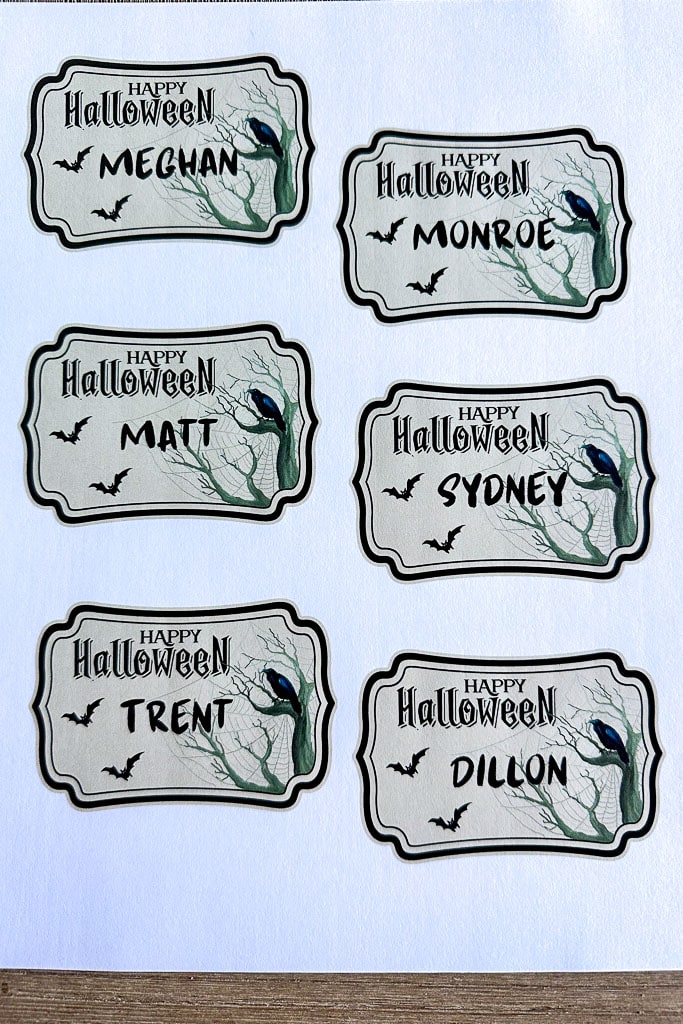 Six Halloween-themed name tags on a wooden surface, each featuring a spooky tree, bats, and a crow. The names on the tags are Meghan, Monroe, Matt, Sydney, Trent, and Dillon. Perfect as DIY Halloween Place Card Holders with "Happy Halloween" at the top of each tag.