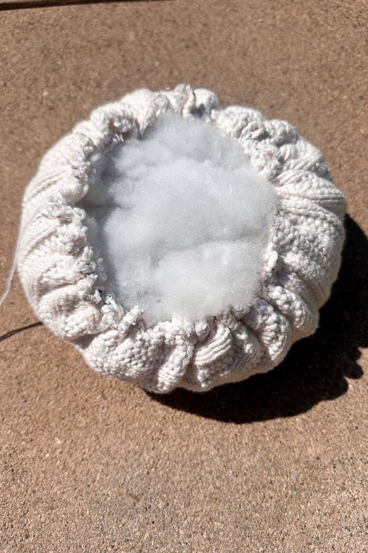 A knitted cream-colored circular object with a gathered edge, partially filled with white stuffing, rests on a textured stone surface in bright sunlight.