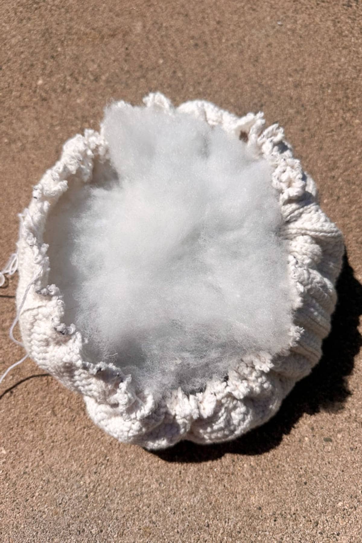 A round, knitted cream-colored pouf placed on a concrete surface, filled with fluffy white stuffing that is visible at the top.