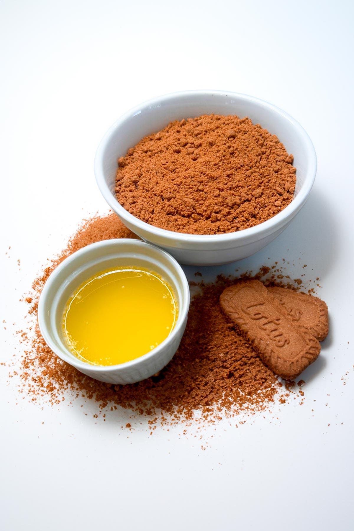 A white bowl filled with crushed Biscoff cookies, a small bowl of melted butter, and a whole Biscoff cookie placed on white background with cookie crumbs scattered around.