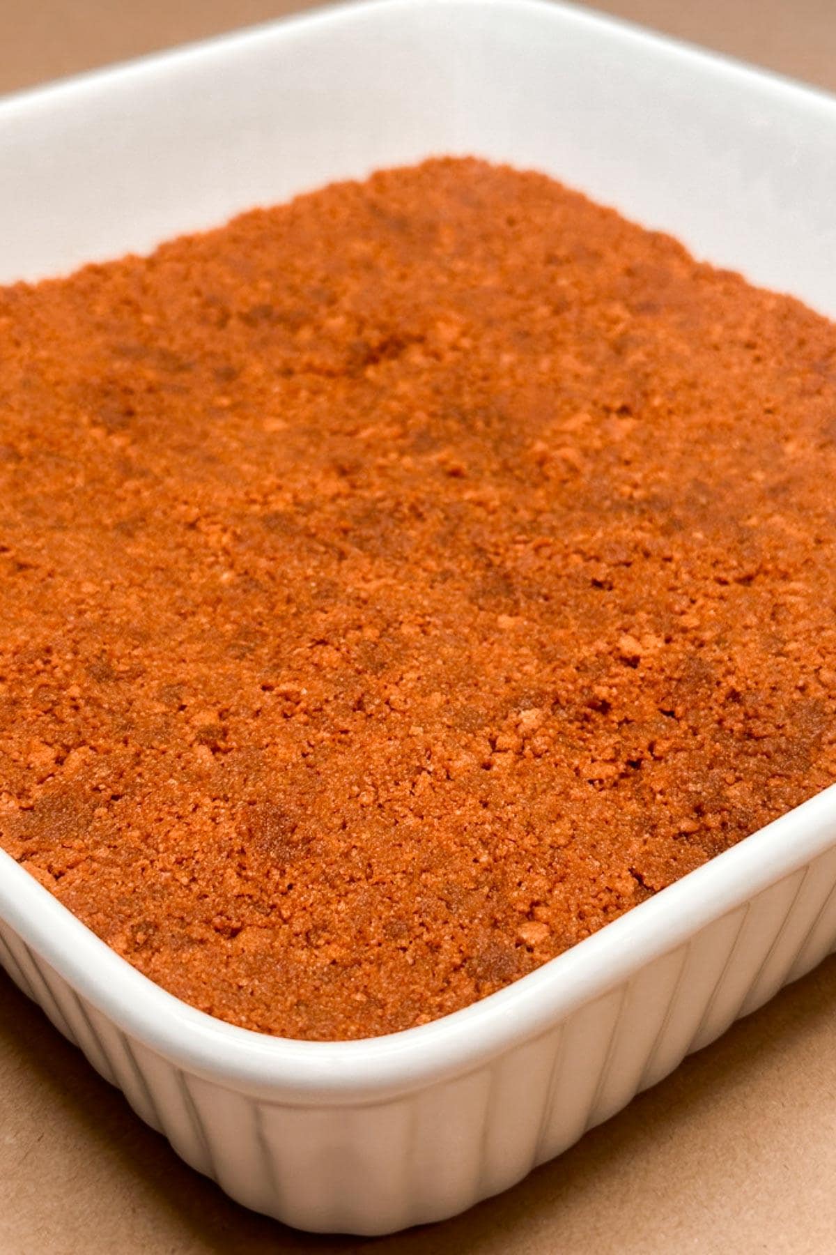 A square white dish filled with a thick layer of finely ground biscoff cookies crushed and pressed into a dish. The surface is evenly spread and has a slightly coarse texture.