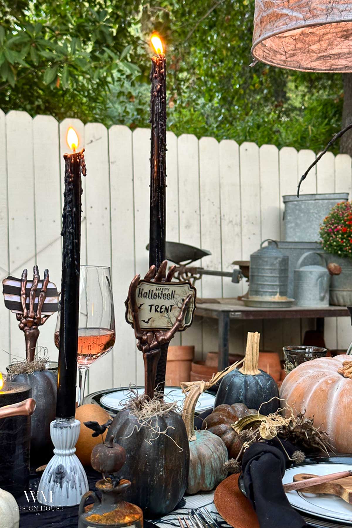 A Halloween-themed table with black candles, skeleton hand holders, assorted pumpkins, a wine glass, and a name card reading “Halloween Trent.” The scene is outdoors, featuring creative DIY Halloween decor set against a wooden fence and garden tools in the background.