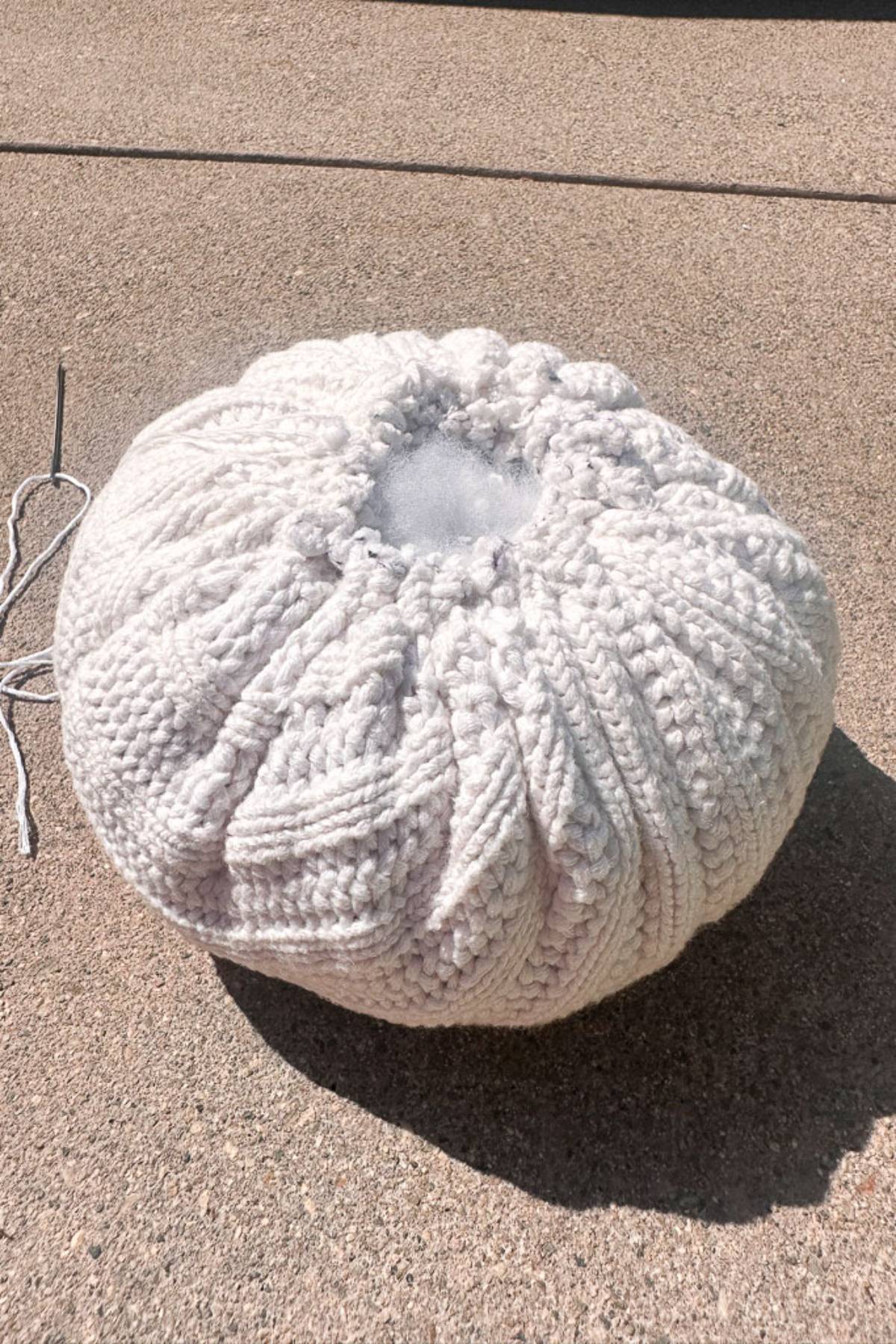 A partially stuffed, round sweater pumpkin lies on a concrete surface. It's made of white yarn with a textured, ribbed pattern, and there's a threading needle with yarn extending from the unfinished opening.