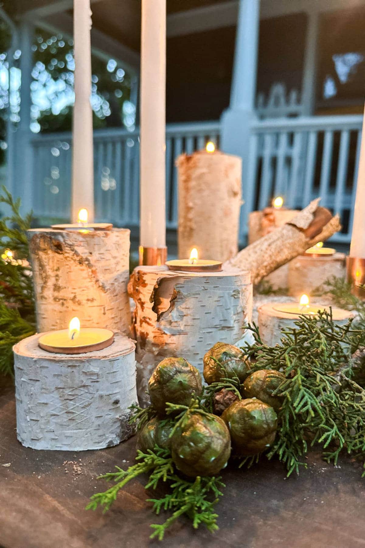 Candles embedded in rustic birch logs are lit on a table, surrounded by greenery and pine cones. The background shows a porch with white railings, creating a cozy outdoor setting.