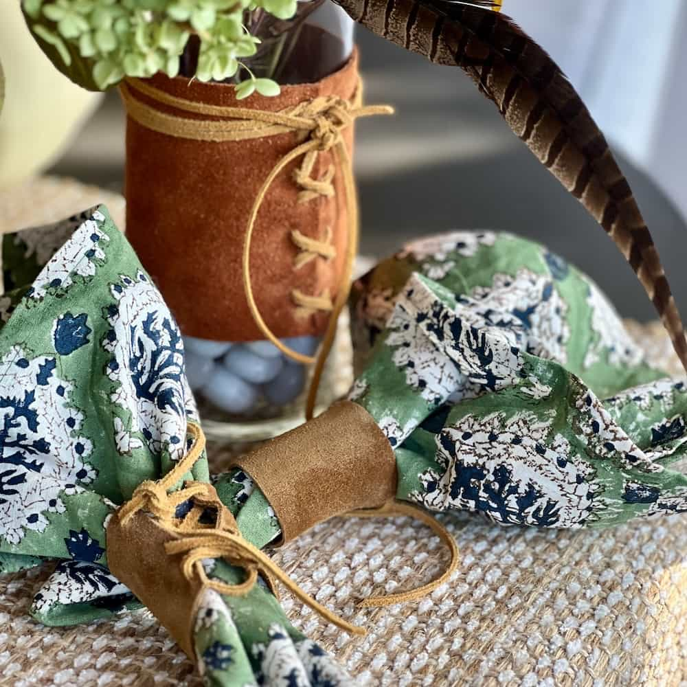 A close-up of two green and white floral fabric napkin holders with brown suede ties, next to a brown suede-covered vase with lace-up detailing, topped with green flowers and a long feather. All items are placed on a woven surface.