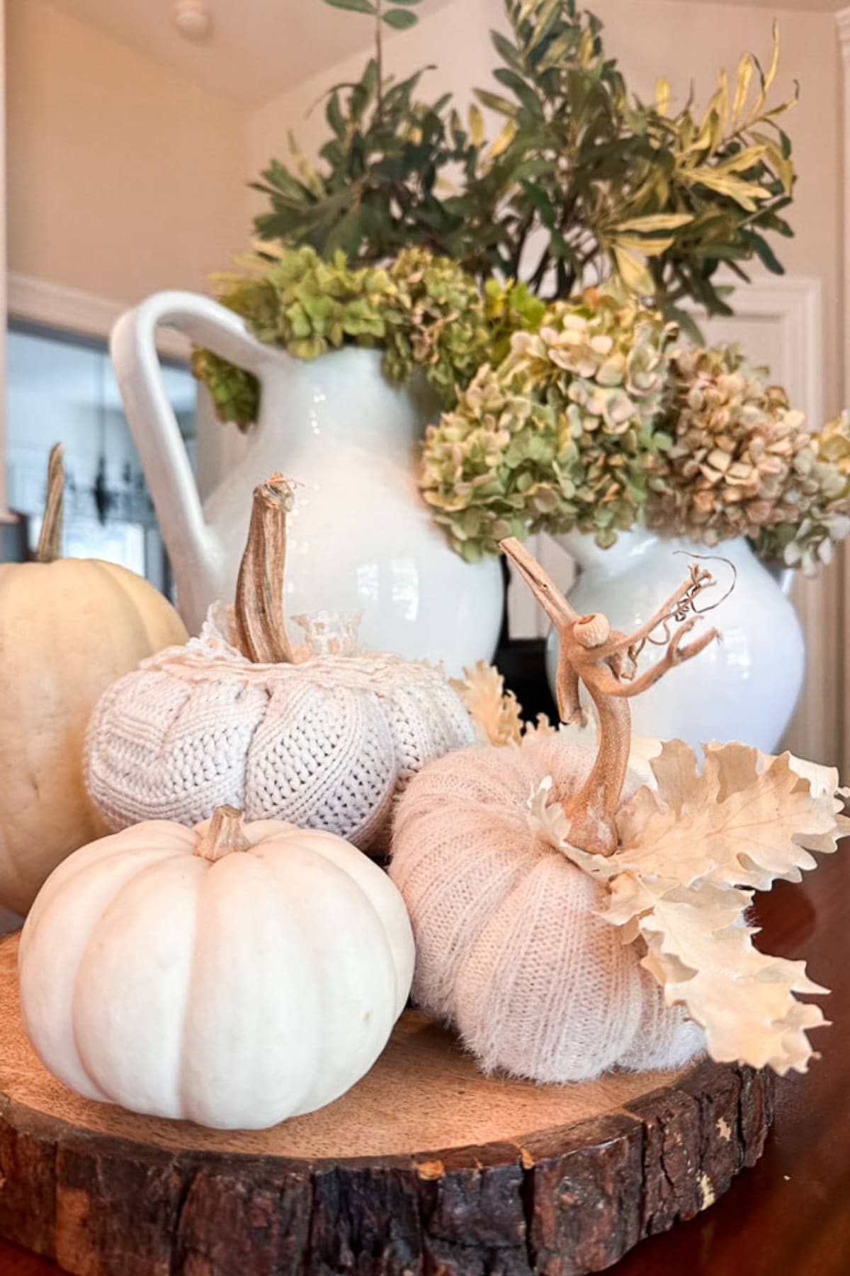 A cozy autumn display featuring white sweater pumpkins arranged on a wooden slice. In the background, there's a white pitcher filled with greenery and dried hydrangeas.