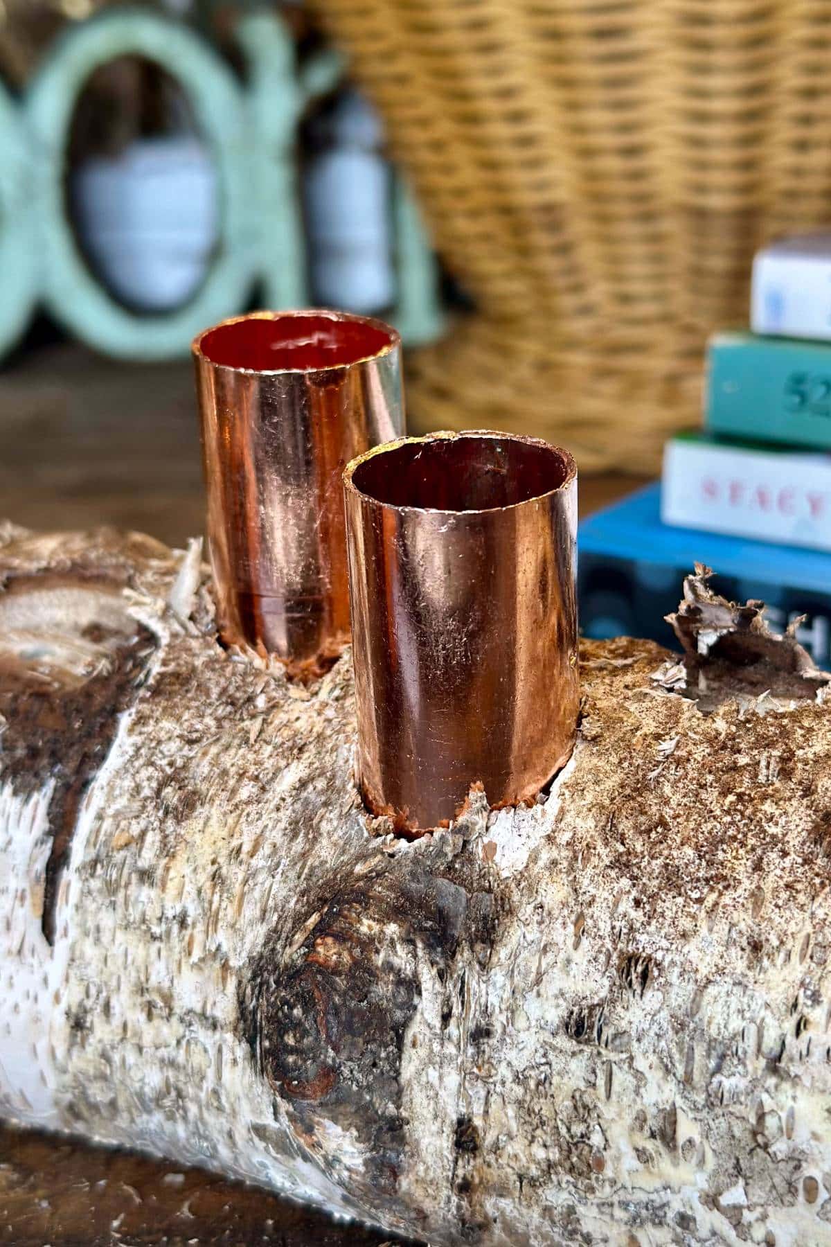 Two copper pipes are embedded vertically into a piece of birch wood. A wicker basket and stacks of books are blurred in the background, adding depth to the rustic setting.