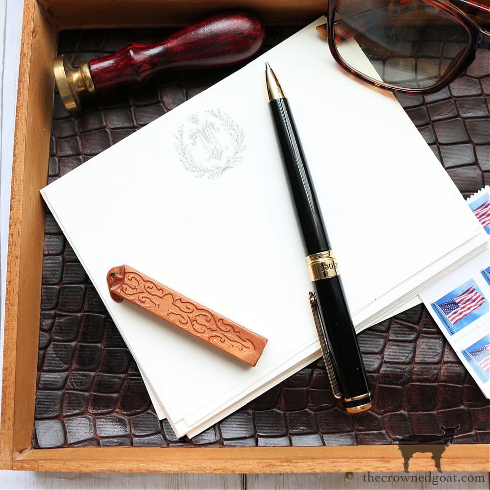 A tray containing a stack of stationery with an embossed emblem, a black and gold pen, a wooden-handled seal, and a pair of sunglasses. Stamps with the US flag are also visible.