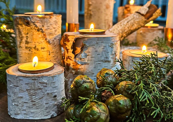 Candles sit on DIY birch wood holders surrounded by evergreen branches and decorative acorns. The warm glow creates a cozy atmosphere.