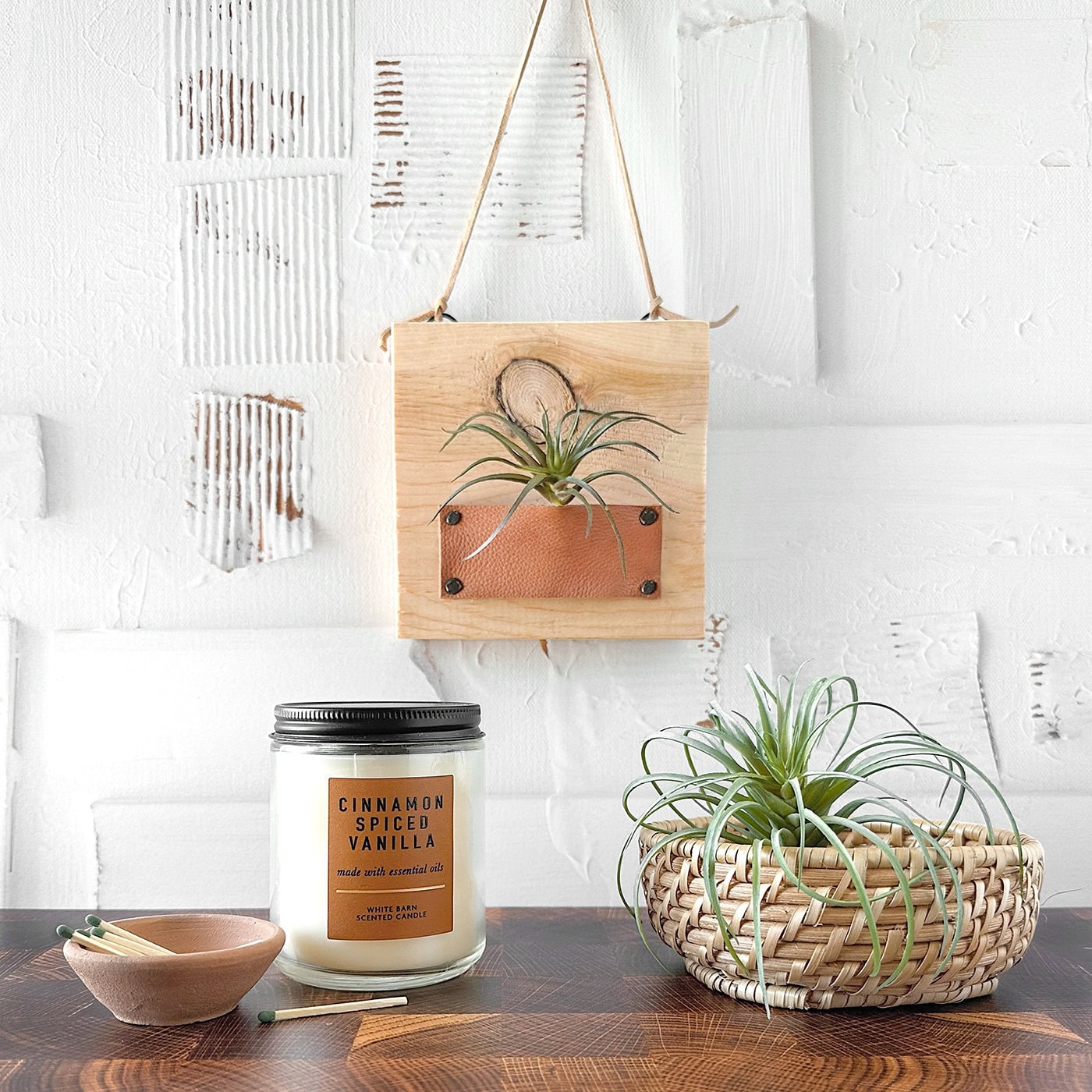 A cinnamon spiced vanilla candle on a wooden table with a small clay dish and matchsticks. A woven basket with an air plant sits nearby. A wooden wall hanging with another air plant is displayed on a textured white wall.