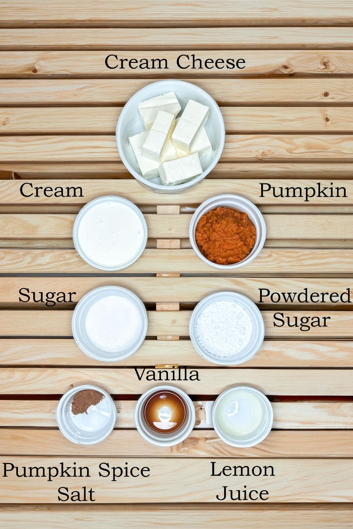 Top view of ingredients on a wooden surface: cream cheese, cream, pumpkin puree, sugar, powdered sugar, vanilla, pumpkin spice with salt, and lemon juice. Each ingredient is labeled in a small bowl.