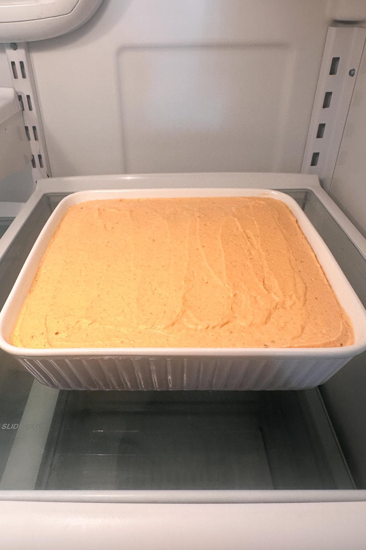 A white rectangular dish filled with a smooth, creamy pumpkin cheesecake mixture, likely a dessert or batter, is placed on a glass shelf inside a refrigerator.