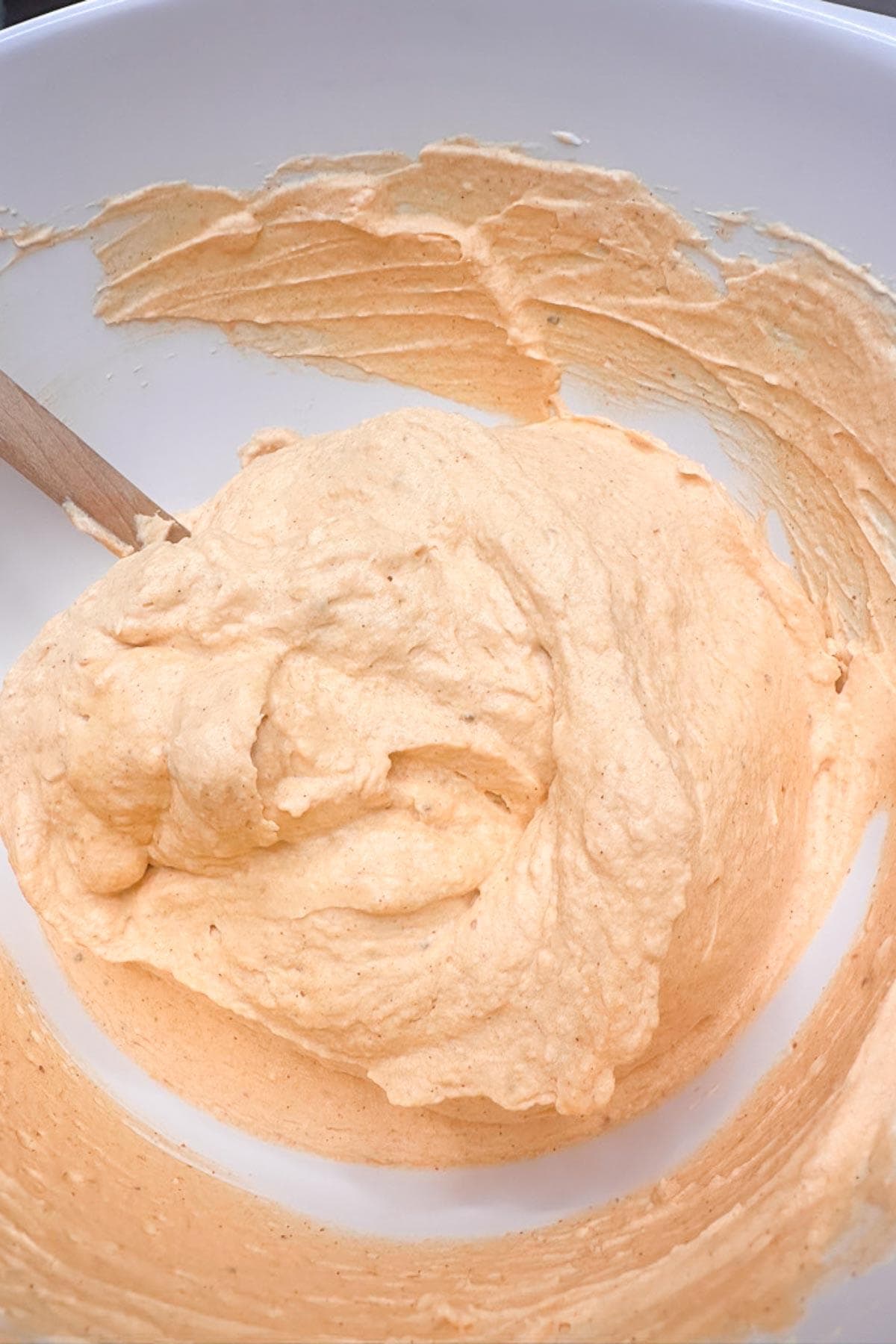 A close-up of a mixing bowl filled with creamy, light orange batter for a pumpkin cheesecake. A wooden spoon is partially visible in the batter, which has a smooth and fluffy texture. The bowl is white, providing a clean background for the mixture.