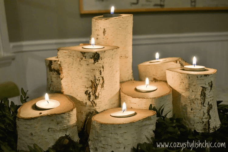 A decorative arrangement of lit tea candles set atop hollowed-out birch logs of varying heights, surrounded by greenery. The logs have a rustic texture, creating a cozy and elegant atmosphere in the room.