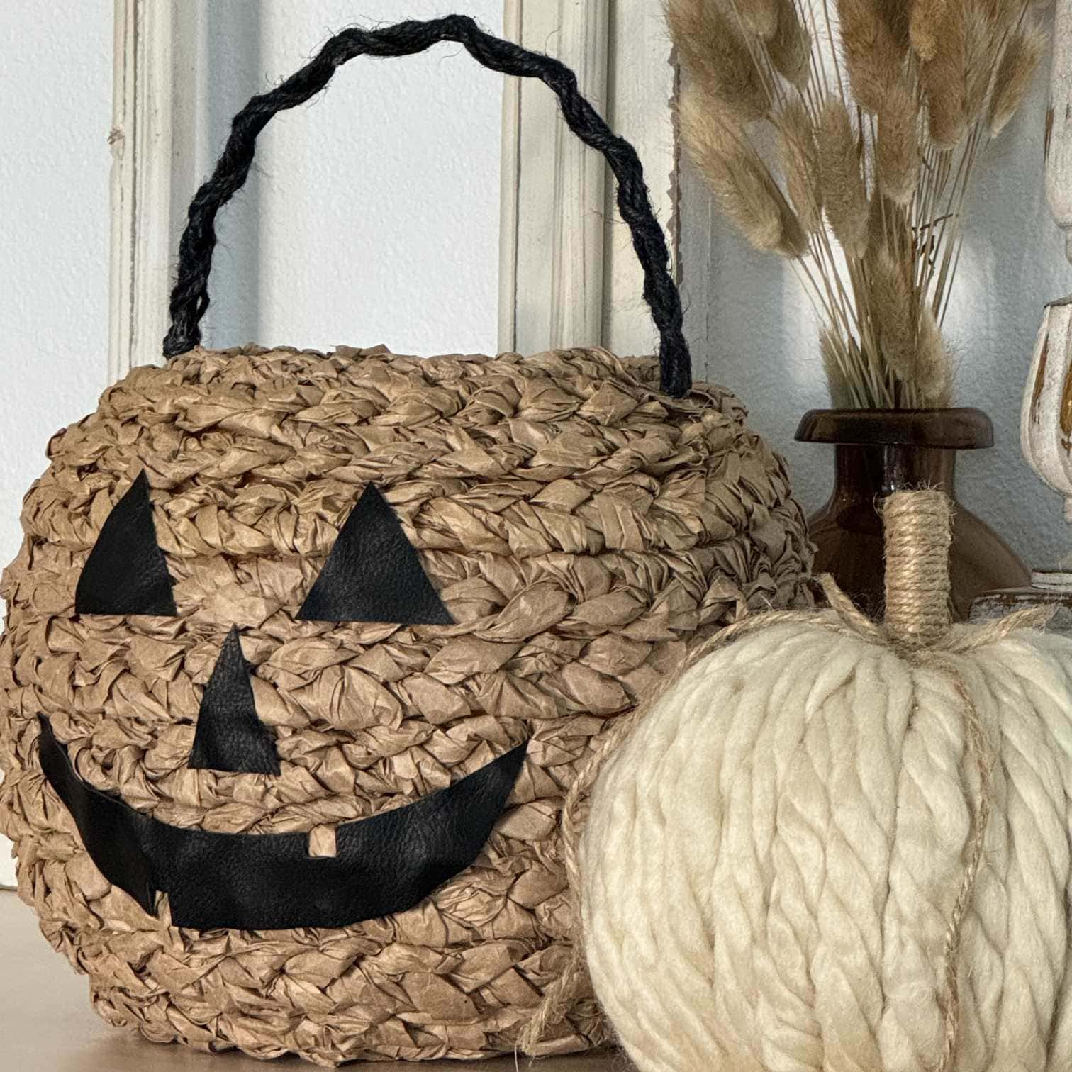 A woven basket with a black jack-o'-lantern face and handle sits next to a cream-colored, yarn-textured pumpkin. In the background, dried decorative grasses are displayed in a brown vase.