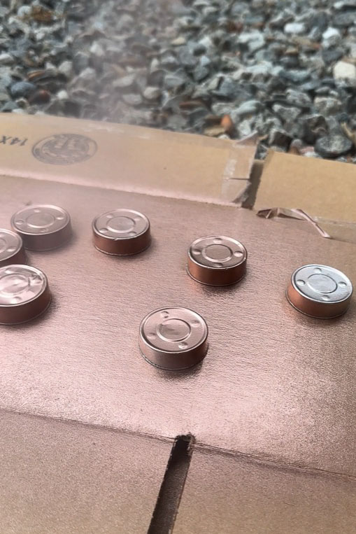 Several small batteries are arranged on a piece of cardboard, resembling the layout of DIY candle holders. The background is composed of gravel.