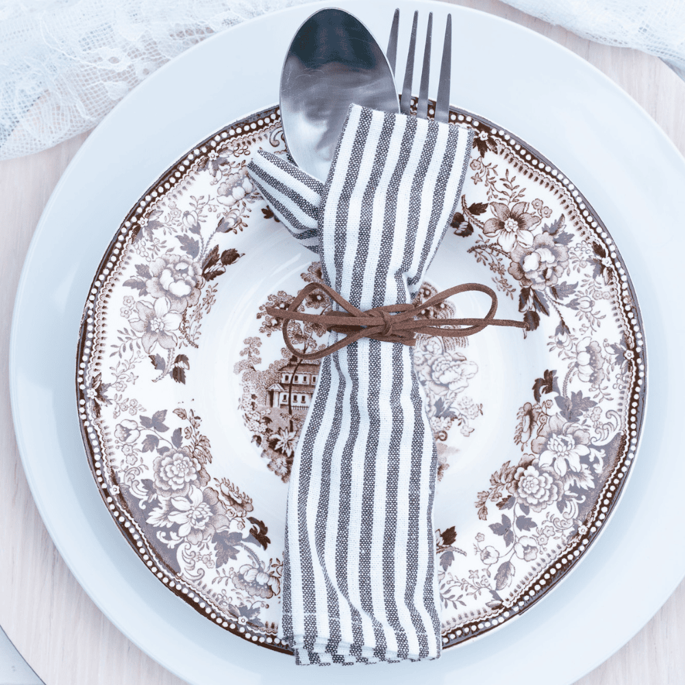 A vintage floral-patterned plate is set on a white plate. A spoon and fork are wrapped in a striped cloth napkin, tied with a brown leather cord, and placed on top of the plates. The setting rests on a light wood surface.