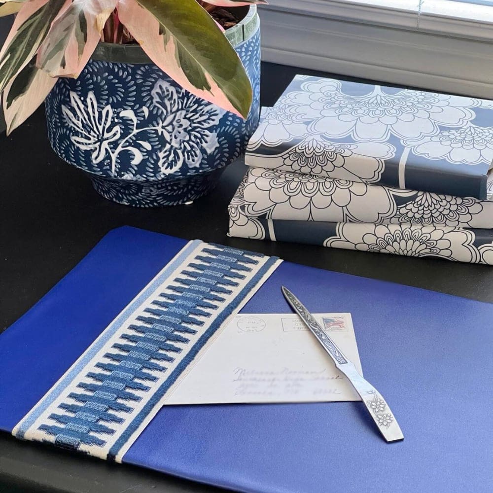 A decorative blue and white plant pot with foliage sits next to a stack of three books wrapped in floral-patterned paper. In the foreground, there's a blue cloth with a striped border, and an envelope with a letter opener on top.