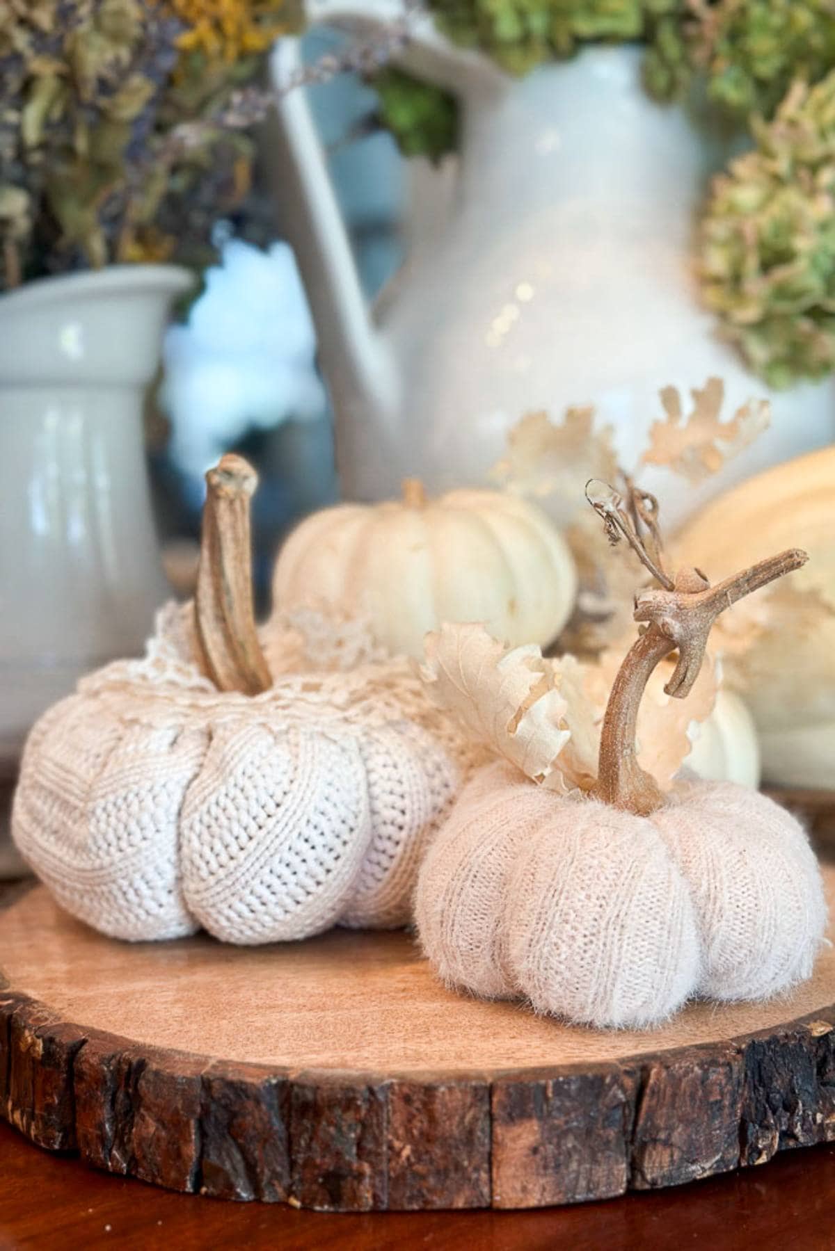 Two decorative sweater pumpkins made of cream-colored sweater fabric sit on a wooden slice. One features lace detailing, and both have realistic stems. In the blurred background, there are other pumpkins and a white pitcher.