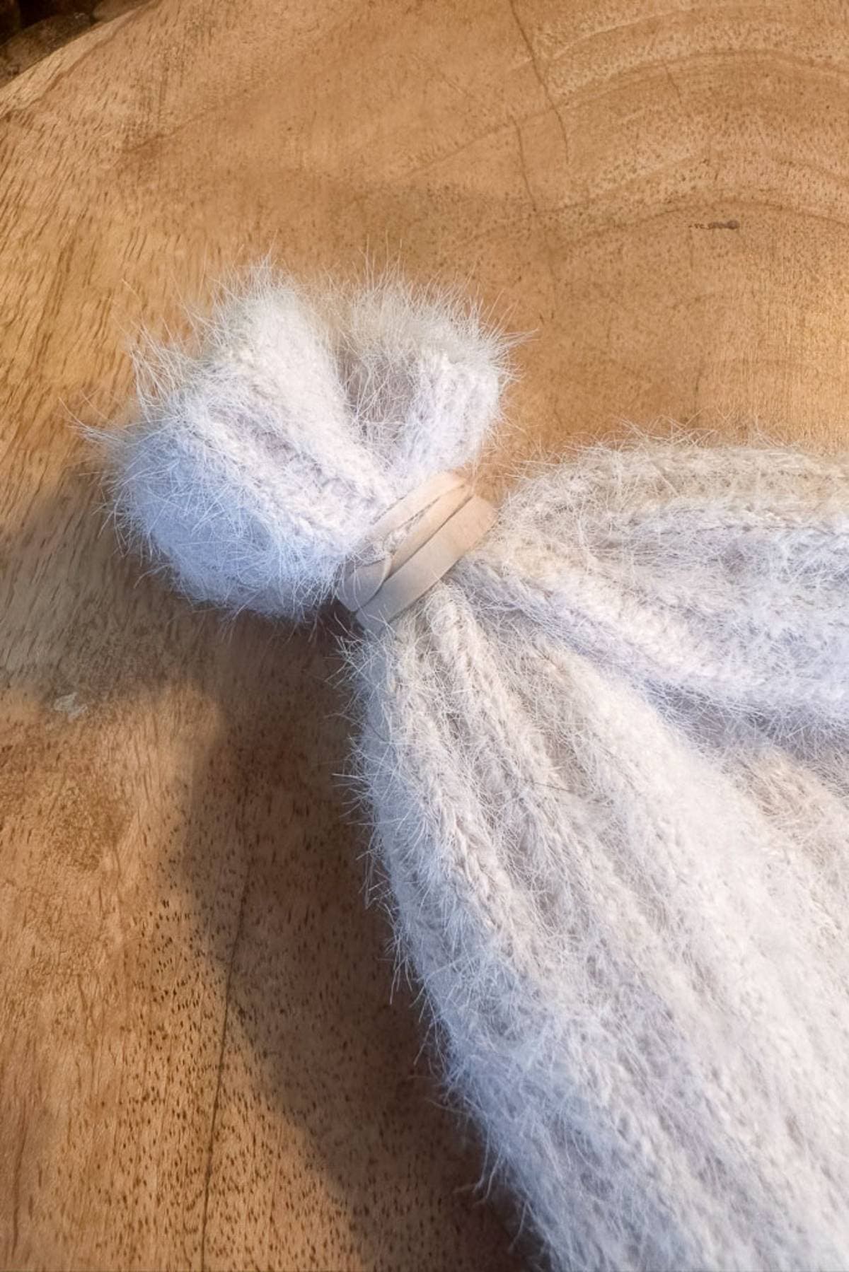 A sweater sleeve that has the end wrapped with a rubber band and lies on a wooden surface. The yarn is light in color, with a fuzzy texture.