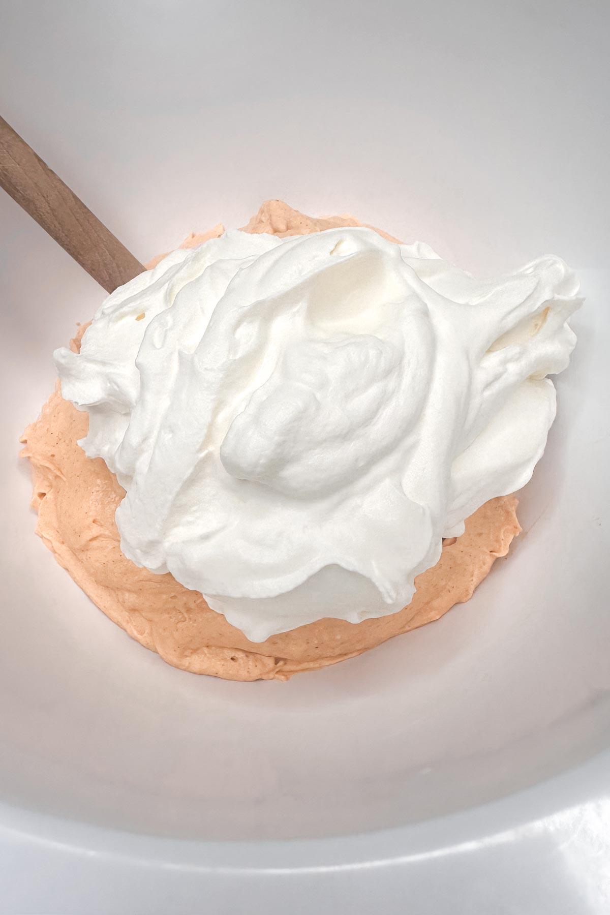 A wooden spoon in a bowl containing a mix of whipped cream and pumpkin cheesecake mixture , likely a dessert or mixture, with visible texture.