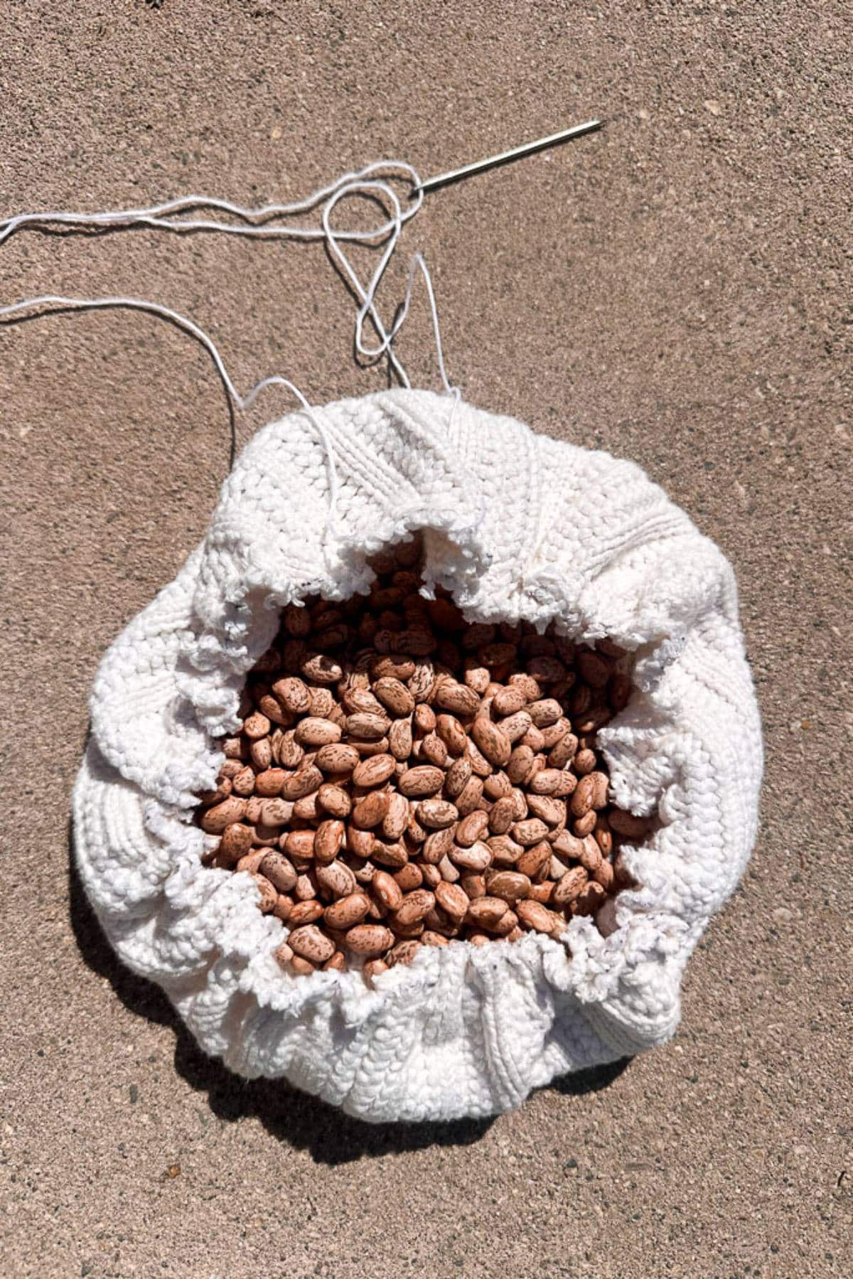 A white knitted pouch, partially open, reveals it is filled with brown beans. A metal knitting needle and some yarn are placed beside it on a textured surface.