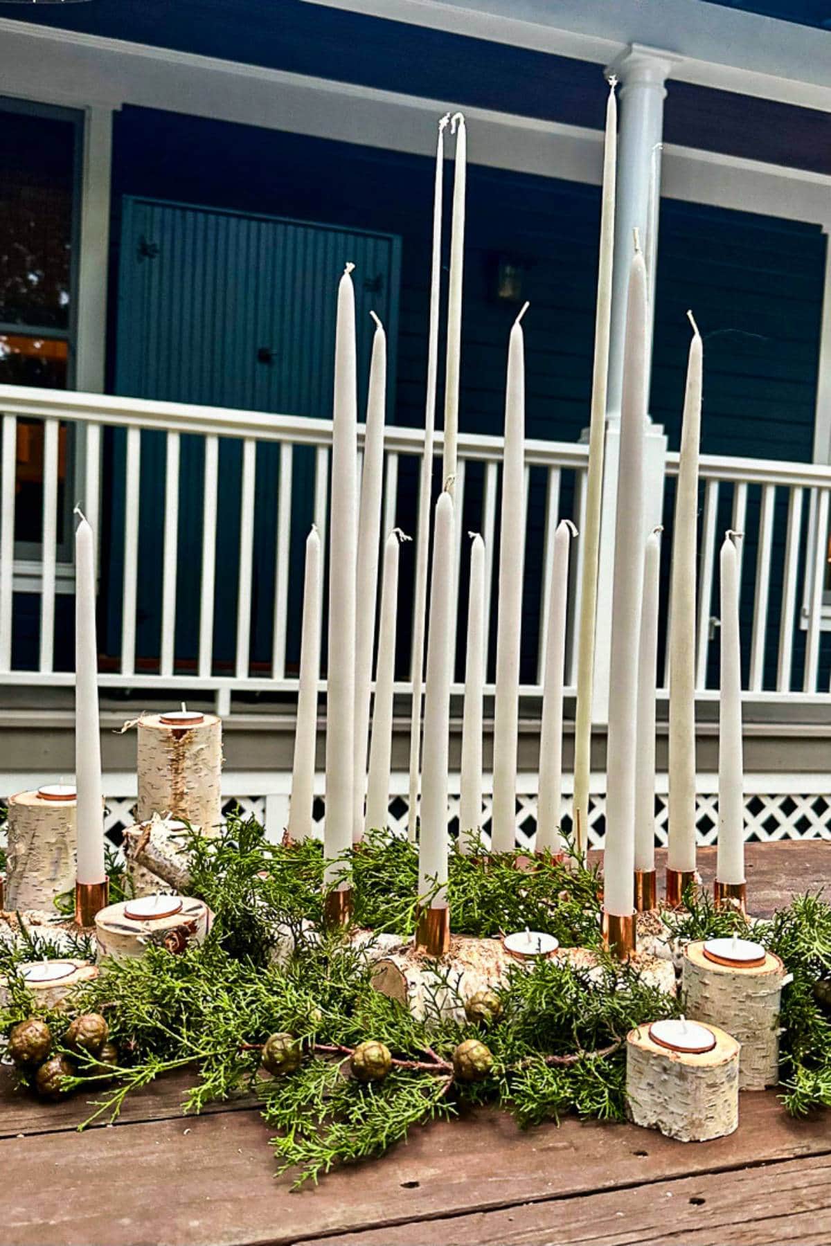 A decorative centerpiece featuring tall, thin white candles on birch holders surrounded by evergreen branches with small pine cones. It is set on a wooden table with a porch and railing in the background.